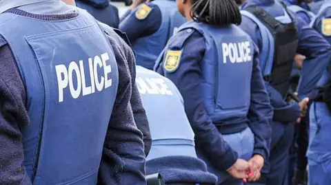 Close up of the backs of police officers standing in formation. The are in uniform and vests with police written on them in white lettering.
