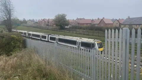 Diana Blamires The train travelling on the route. In front is a silver metal fence, and behind it is a housing estate.