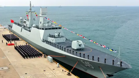 VCG via Getty Images The large naval ship is pictured at dock with flag bunting and crowds of military officers lining the doc at the unvailing ceremony, against a blue sea and horizon.