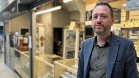 Mark Wilson is standing in front of glass counters with watches at his stall. He has short brown hair, which is combed back, and a beard. He is wearing a khaki shirt and a dark grey jacket.