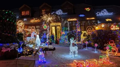 A house with Christmas lights covering the roof, walls and most of the front garden with small lights dotted over trees and hedges and larger lights in the shape of snowmen and reindeers located across the grass. 