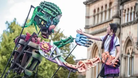 A large puppet shakes hands with a woman in a street. The woman, on the right of the image, has black and purple hair and is wearing a white shirt and a dark skirt.