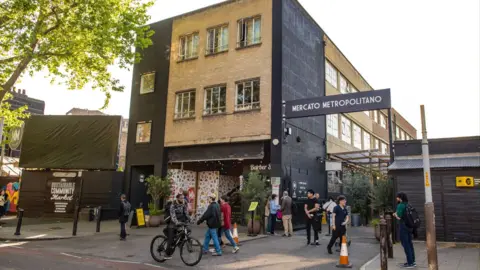 A lively urban street scene featuring Mercato Metropolitano’s entrance, brick building facade, greenery, signage, and people walking and cycling nearby.