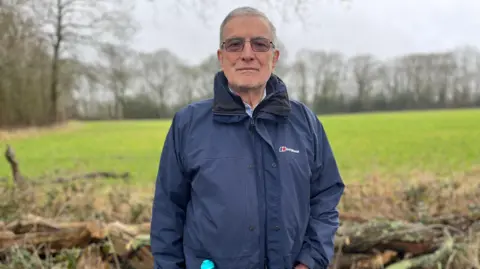 Julia Gregory/BBC Chairman of the Normandy Action Group, Mike Aaronson. Mike is stood at the prospective site. He is wearing a navy mac coat and light sensitive glasses. 