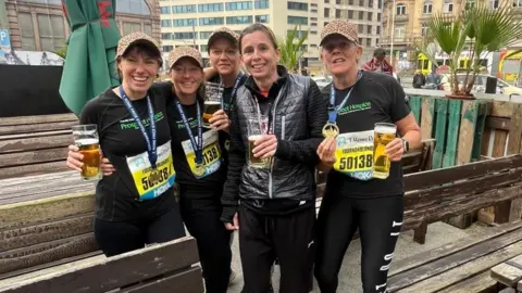 Prospect House The five women, all smiling to camera, in an outside bar holding pint glasses, and still wearing their black marathon gear, including race bibs and medals