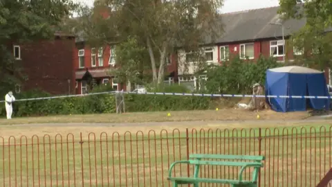 A forensic officer in white protective clothing is standing on a grassed area, with a tent near to some fencing.