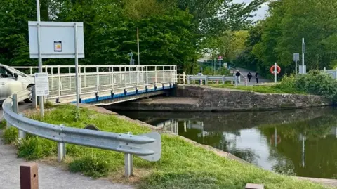 Photo taken from carpark near to the canal. A bridge with a silver car driving on top of it is seen stretching along the width of the canal. 