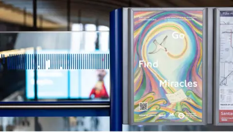 TFL Inside a blue station display board, the Go Find Miracles poster shows a white bird having flown to form a pattern of swirls coloured purple, pink, yellow and blue