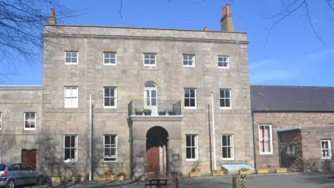 The States of Alderney building is three storeys and made from grey brick. There is a covered archway at the front of the building with a balcony at the top.