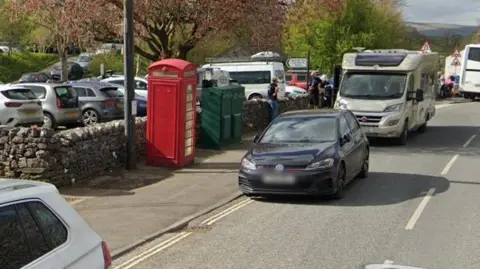 A traditional red phone box next to a busy car park with cars, vans, a motorhome and a coach.