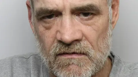 Warren Spence is looking at the camera in this police mugshot. He has short grey hair which is receding and a grey beard. He is wearing a grey top.