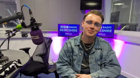 Tommy Longhurst is sat in one of BBC Radio Berkshire's studios. He is wearing a denim jacket and two silver necklaces. He is wearing blue sunglasses and has hair brushed back.