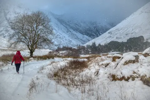 Brian Colston A walker in a red jacket going along a snowy path, with mountains in the distance and snow covering the area