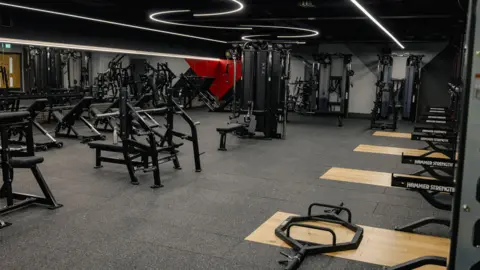 A gym room with squat racks on one wall and bench presses in front of a mirror wall.