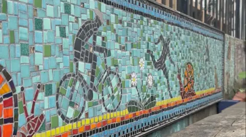 Nabiha Ahmed A mosaic is featured behind a wooden bench. The colours are blue, yellow, orange and black and feature figures running and cycling along the canal, next to plants and snails