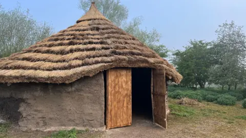 TOM JACKSON/BBC A circular building with a wall made from grey clay, with a wooden door ajar at the front. There is a thatched roof made from several layers of thatching leading up to a single point. Behind it there are trees and shrubs. 