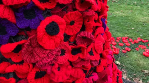 A cascade of knitted poppies around a tree trunk meets the reused plastic bottles on the ground in Queen's Park, Loughborough