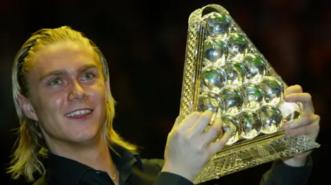 Getty Images Paul Hunter lifts The Masters trophy. The trophy is a glass figurine of a triangle of red balls. Hunter has blond braided hair.