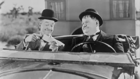 Getty Images A black and white photo of two men sitting in a car with both wearing black bowler hats