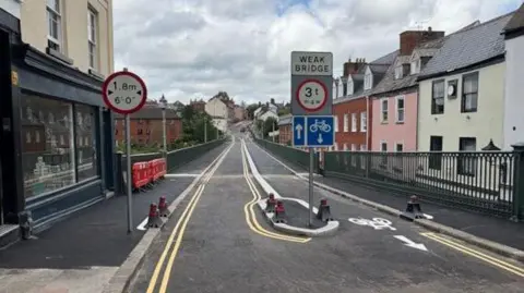 A narrow bridge with road signs including a 1.8-meter (6 feet) height restriction on the left, a central warning for a weak bridge with a 3-ton weight limit, and a blue sign allowing bicycles to proceed straight. The road has yellow lane markings and bollards guiding traffic. Red barriers are visible on the left sidewalk, and buildings line both sides of the bridge.