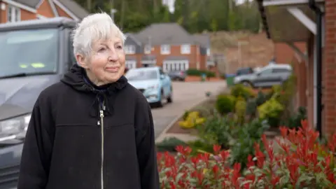 Irene Taylor is standing outside a home with a van and car on the left and plants outside the row of houses on the right