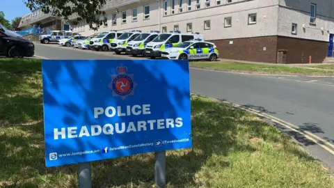 Police Headquarters blue sign with white bold letters. Row of police cars and vans behind the dign, in front of the police building.