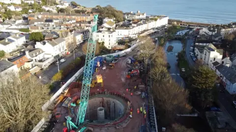 A view from above of a coastal town, with the sea in the distance. In the foreground is a building site - with a large blue crane - where a large circular concrete tank is being built in the ground