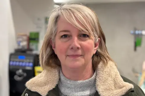 BBC/Mark McAlindon A head-and-shoulders image of Sandy Lancaster - a woman with blonde hair in a long bob. She is wearing a khaki-coloured coat with a cream faux fur collar and she is smiling and looking directly at the camera. She is standing in her sandwich shop.