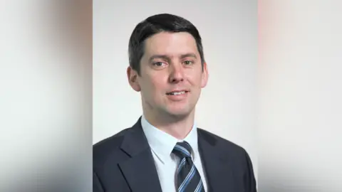 Suffolk County Council A portrait photo of Chris Chambers. He smiles at the camera. He has short black hair and wears a navy suit with a white shirt and a black, blue and white tie.