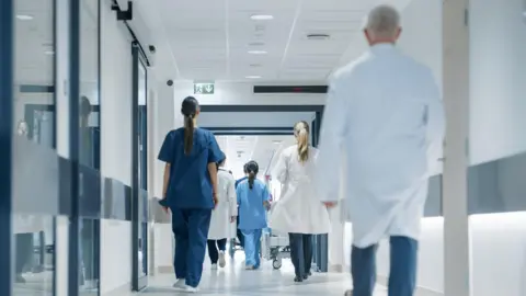 Hospital hallway with doctors, nurses and specialists with their back to camera. They are in a variety of hospital uniforms - dark and light blue scrubs, white coats.