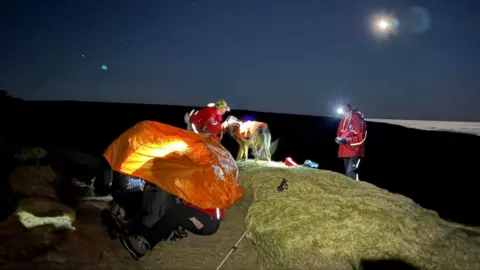 People warming in an orange shelter tent on a mountain with two Edale Mountain Rescue Team volunteers and their dog nearby on a winter night