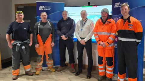 Jersey Electricity Jersey Electricity engineers are standing in front of a screen and two large posters. There are six men. 