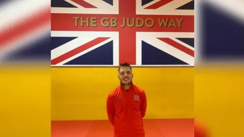 Rowan Kinsella Mr Kinsella stands in front of a Union Jack which has 'THE GB JUDO WAY' written on it. He is standing with his arms behind his back and is smiling at the camera. He is wearing a red, long-sleeve jumper with the Great Britain team logo on it.