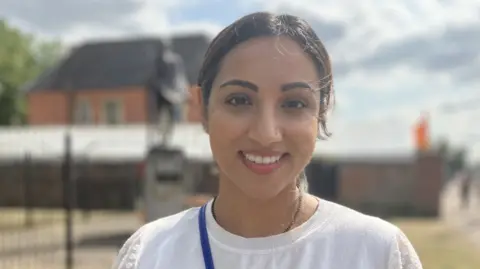 A woman wearing a white T-shirt smiles towards the camera. Behind her is an out-of-focus building