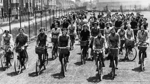 BPEOSA collection A large group of women, in a black and white photo, riding bikes on a stretch of land with houses and a fence to one side. They are all wearing coats and hats and are smiling and looking at the camera. 