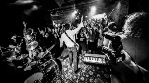 Veronika Zolotoverkha A black and white photo taken of a band from behind as they perform on stage. The audiences if full or predominantly young women with their hands in the air