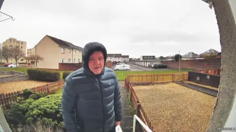 A man in a hooded jacket on the front doorstep of a house, with the image from a doorbell security camera. Several other homes and cars are visible in the background.