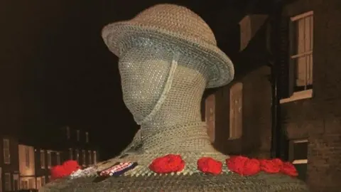 The Secret Yarn Bomber A knitted head of a soldier is on top of a post box. It is beige in colour and there are red poppies at its base.