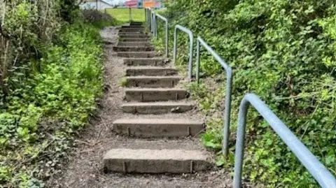 An outdoor footpath made up of a series of concrete steps running up a small incline towards a semi-urban area at the top. On both sides of the steps, there are metal handrails painted grey, running parallel to the path. Green vegetation lines both sides of the path.