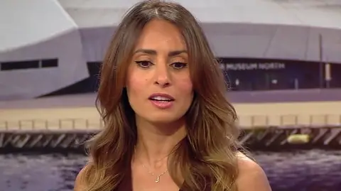 Layla Hamood with long brown hair and wearing a brown sleeveless knitted top with a necklace with the initial J in the BBC Breakfast studio with Salford Quays in the background.