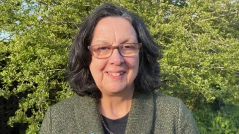 CPRE A mid-shot of Sarah standing outside in sunshine, smiling to camera. She is an older lady with glasses and greying dark hair and is wearing a green tweed-style jacket. There is a tree in leaf just behind her.