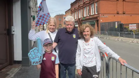 Two women, a man and a boy gather together on a pavement next to a barrier. One of the women has a flag above her head. The boy has a foam hand.