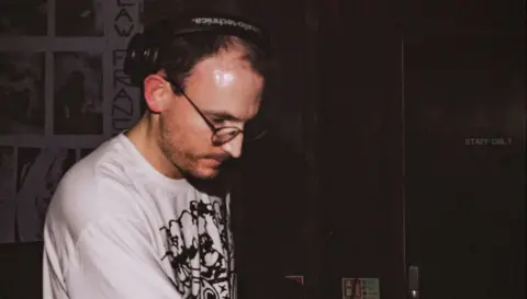 Strange Brew Leigh Dennis is standing in a dark room wearing a white T-shirt and black headphones. He is looking down onto a desk.