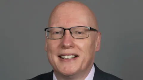 NHS A man with a clean-shaven head and metal-framed glasses smiles at the camera against a grey background in a corporate headshot.