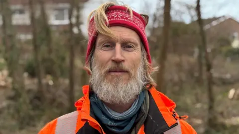 A man with a thick grey beard and long, blond hair smiles directly into the camera. He is wearing a red and white patterned headband and a bright orange high visibility jacket, with a badge of a tree on the left side of his chest. He is standing in front of a wood with wood chippings in the background.