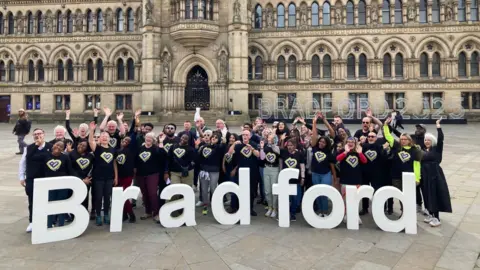 Fifty people standing in front of polystyrene letters spelling out Bradford with City Hall in the background