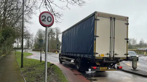 A new 20mph sign in Castle Douglas, A blue-sided lorry is driving past on to a roundabout. It is a miserable, wet day. 
