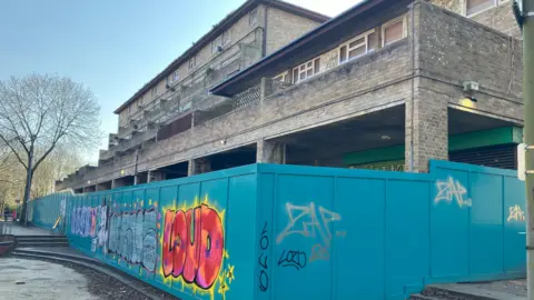 Amy Holmes/BBC A long teal construction barrier covered in graffiti runs alongside a stepped walkway in front of a large, weathered concrete building with multiple levels and balconies.