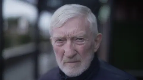 Brendan Hughes mira directamente a la cámara. Tiene el pelo gris y una barba corta con una mezcla de verde y marrón en los ojos. Lleva un abrigo azul oscuro. El fondo de la imagen es borrosa, pero se ve un poco de vegetación y un gran marco de metal. 