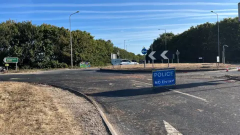 Clare Worden/BBC. Un cartel azul de POLICÍA - PROHIBIDO EL PASO se encuentra en una rotonda. Hay un coche blanco al otro lado de la rotonda.